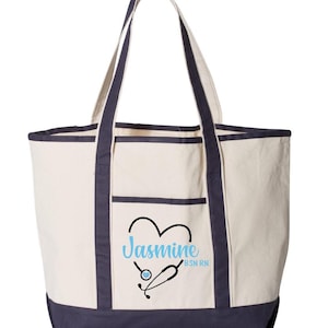 May include: Cream-colored tote bag with navy blue trim and handles. The bag features a front pocket and a design of a heart with a stethoscope and the name "Jasmine" in light blue, with "BSN RN" below.