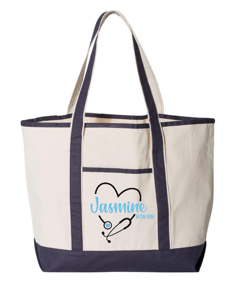 May include: Cream-colored tote bag with navy blue trim and handles. The bag features a front pocket and a design of a heart with a stethoscope and the name "Jasmine" in light blue, with "BSN RN" below.