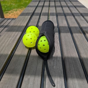 Peut inclure: Trois balles de pickleball jaune fluo avec de multiples trous, &agrave; c&ocirc;t&eacute; d'un &eacute;tui noir zipp&eacute;. L'&eacute;tui est ouvert, r&eacute;v&eacute;lant une balle &agrave; l'int&eacute;rieur. Les articles sont sur une table de pique-nique en bois.