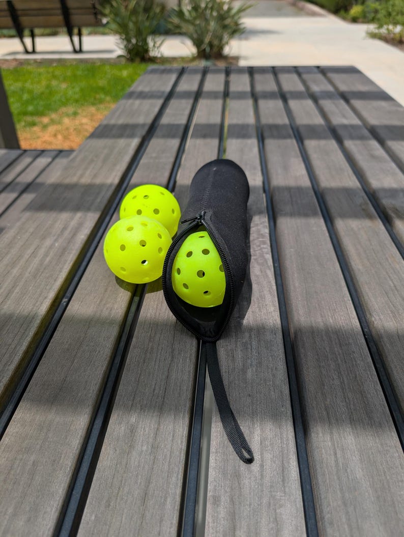 Peut inclure: Trois balles de pickleball jaune fluo avec de multiples trous, &agrave; c&ocirc;t&eacute; d'un &eacute;tui noir zipp&eacute;. L'&eacute;tui est ouvert, r&eacute;v&eacute;lant une balle &agrave; l'int&eacute;rieur. Les articles sont sur une table de pique-nique en bois.