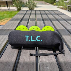 Peut inclure: Un &eacute;tui en n&eacute;opr&egrave;ne noir avec le texte "T.L.C." en turquoise, contenant trois balles de pickleball vert n&eacute;on. L'&eacute;tui a une fermeture &eacute;clair et une petite sangle noire. Les balles ne sont pas incluses.