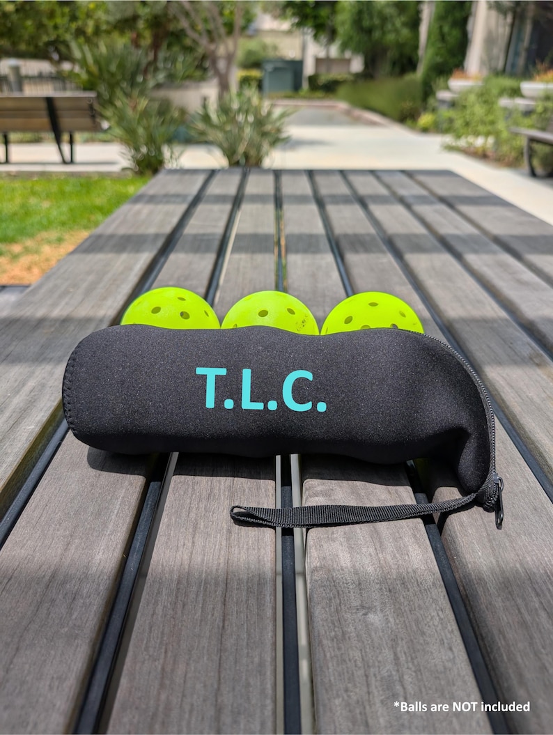 Peut inclure: Un &eacute;tui en n&eacute;opr&egrave;ne noir avec le texte "T.L.C." en turquoise, contenant trois balles de pickleball vert n&eacute;on. L'&eacute;tui a une fermeture &eacute;clair et une petite sangle noire. Les balles ne sont pas incluses.