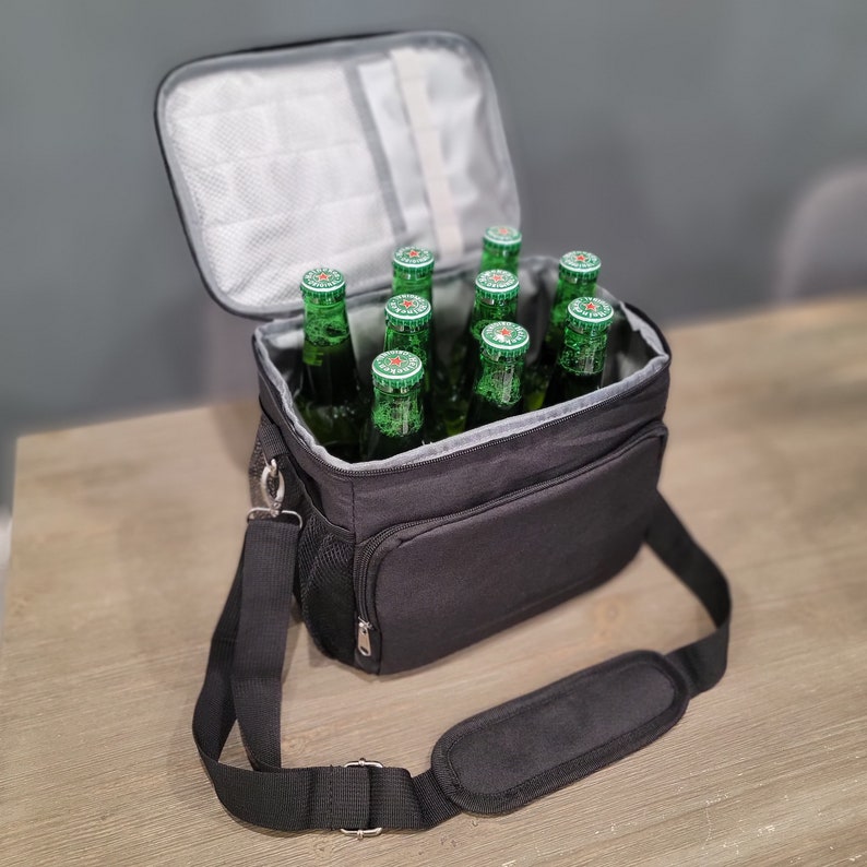 May include: A black insulated cooler bag with a shoulder strap, open to reveal several green glass beer bottles. The cooler has a zippered front pocket and a mesh side pocket. The interior is lined with a silver material.