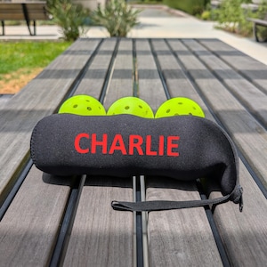 Peut inclure: Un &eacute;tui de pickleball en n&eacute;opr&egrave;ne noir avec le nom "CHARLIE" en lettres rouges. Trois balles de pickleball vert fluo sont visibles. L'&eacute;tui a une fermeture &eacute;clair noire et une petite poign&eacute;e noire. L'image est prise &agrave; l'ext&eacute;rieur sur une table en bois.