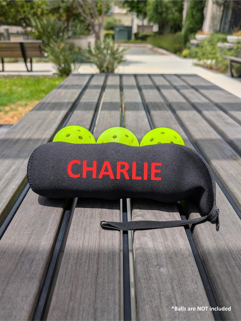 Peut inclure: Un &eacute;tui de pickleball en n&eacute;opr&egrave;ne noir avec le nom "CHARLIE" en lettres rouges. Trois balles de pickleball vert fluo sont visibles. L'&eacute;tui a une fermeture &eacute;clair noire et une petite poign&eacute;e noire. L'image est prise &agrave; l'ext&eacute;rieur sur une table en bois.