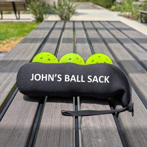 Peut inclure: Sac de balles en n&eacute;opr&egrave;ne noir avec le texte "JOHN'S BALL SACK" en blanc. Trois balles de pickleball vert fluo sont partiellement visibles. Le sac a une petite poign&eacute;e noire et repose sur une table en bois.