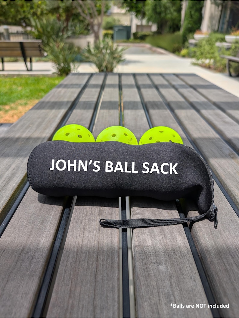 Peut inclure: Sac de balles en n&eacute;opr&egrave;ne noir avec le texte "JOHN'S BALL SACK" en blanc. Trois balles de pickleball vert fluo sont partiellement visibles. Le sac a une petite poign&eacute;e noire et repose sur une table en bois.