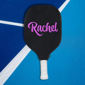 May include: A black pickleball paddle with a white handle and a pink "Rachel" name on the face.