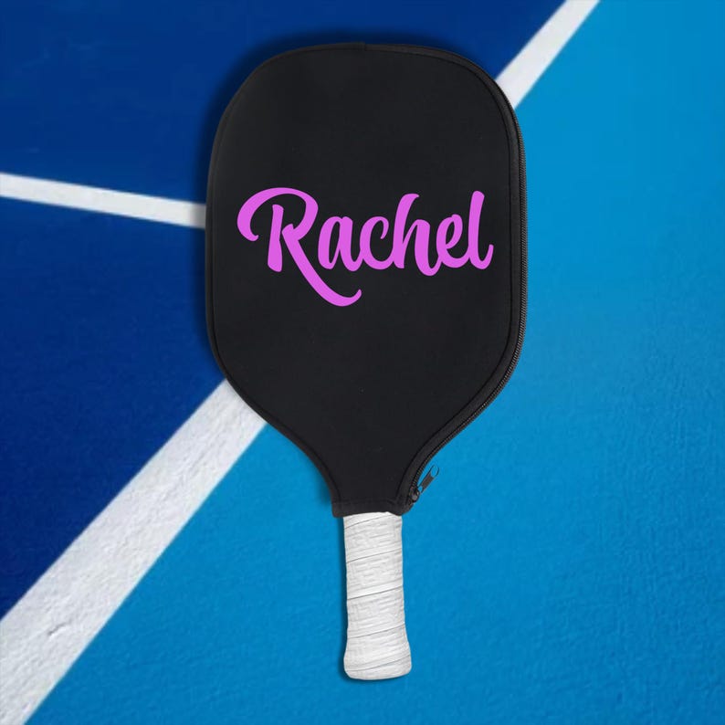 May include: A black pickleball paddle with a white handle and a pink "Rachel" name on the face.
