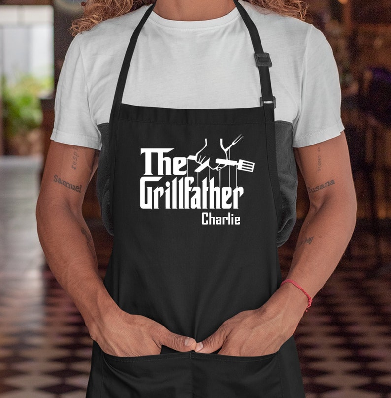 May include: Black apron with white text that reads "The Grillfather Charlie". The apron has a white graphic of a hand holding a spatula and a fork.