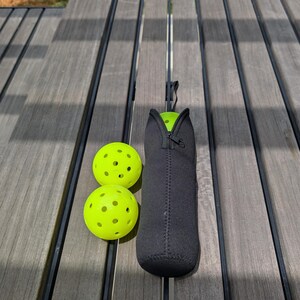 Peut inclure: Deux balles de pickleball jaune fluo avec plusieurs trous et un &eacute;tui de transport en n&eacute;opr&egrave;ne noir avec une fermeture &eacute;clair. L'&eacute;tui est con&ccedil;u pour contenir une balle de pickleball. Les articles sont sur une surface en bois grise.