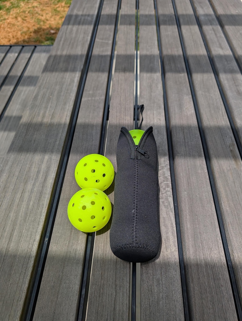 Peut inclure: Deux balles de pickleball jaune fluo avec plusieurs trous et un &eacute;tui de transport en n&eacute;opr&egrave;ne noir avec une fermeture &eacute;clair. L'&eacute;tui est con&ccedil;u pour contenir une balle de pickleball. Les articles sont sur une surface en bois grise.