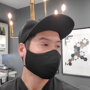 May include: A person wearing a black face mask and a black baseball cap. The mask is made of a soft, breathable fabric and has a comfortable fit.