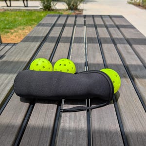 Peut inclure: Trois balles de pickleball vert fluo &agrave; fossettes noires reposent sur une table en bois. Un &eacute;tui noir zipp&eacute; contient une balle, avec deux autres visibles. La table a un aspect vieilli, avec un cadre de parc en arri&egrave;re-plan.