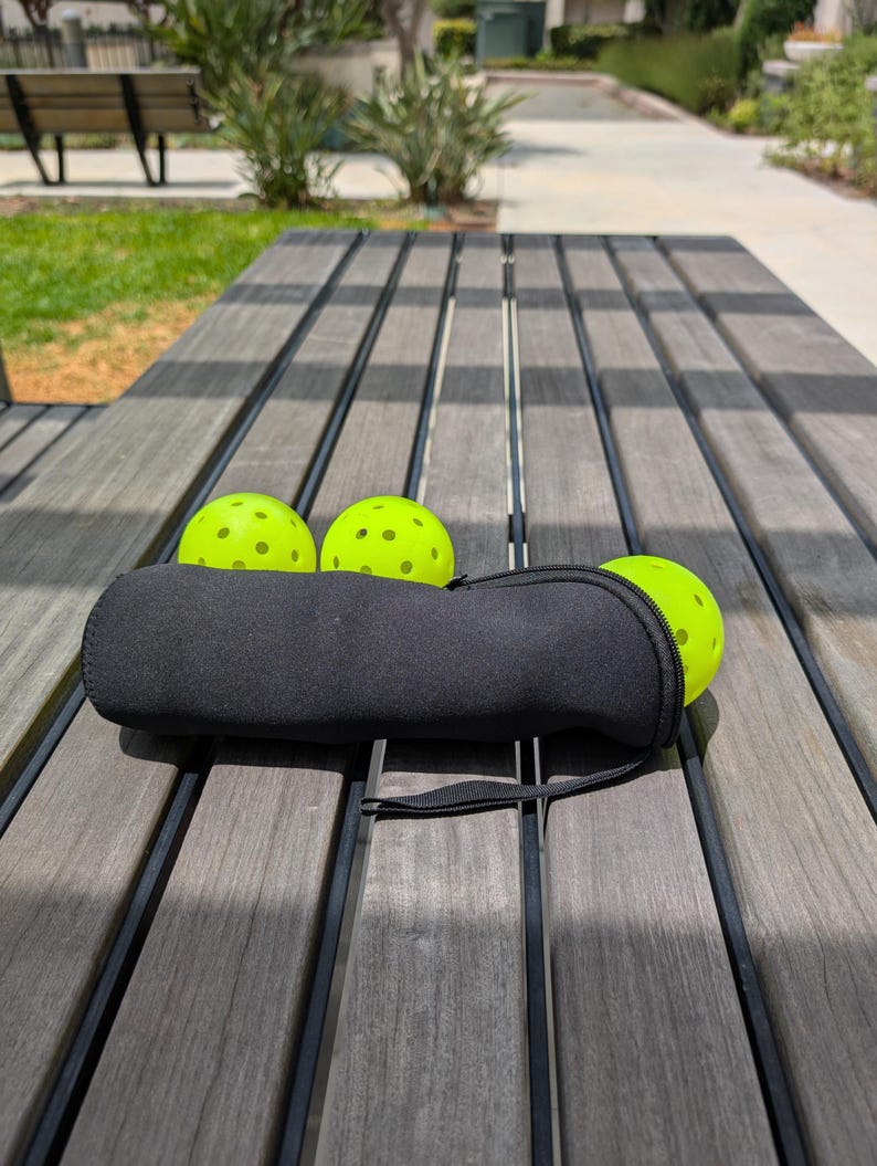 Peut inclure: Trois balles de pickleball vert fluo &agrave; fossettes noires reposent sur une table en bois. Un &eacute;tui noir zipp&eacute; contient une balle, avec deux autres visibles. La table a un aspect vieilli, avec un cadre de parc en arri&egrave;re-plan.