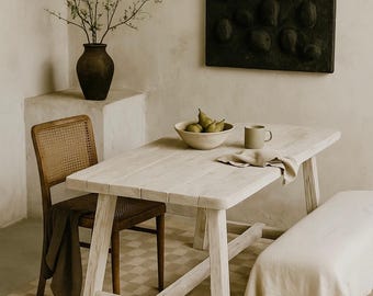 Puede incluir: Una mesa de comedor de madera clara con base de caballete, acompañada de una silla con respaldo de rejilla y un textil oscuro, un banco cubierto con una tela clara, un cuenco de cerámica con peras verdes, una taza y un jarrón oscuro con ramas.
