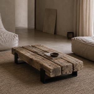 Peut inclure: Une table basse composée de trois poutres en bois épais et patiné à la surface rugueuse et texturée. La table est soutenue par deux pieds en métal noir en forme de U et présente un petit bol marron foncé posé sur un objet plat.