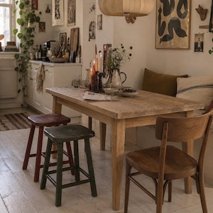May include: A rectangular wooden dining table with a natural finish, accompanied by a dark red stool, a dark green stool, and a wooden chair with a curved backrest. The table surface holds a candle holder with lit candles, a pitcher, and a newspaper.