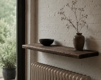 May include: A dark brown wooden floating shelf mounted on a white brick wall. On the shelf sits a small black bowl and a brown ceramic vase containing a thin, delicate branch with tiny buds. Below the shelf is a white vertical radiator.
