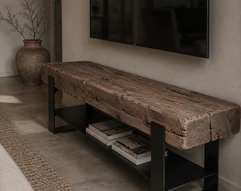 Puede incluir: Un banco rectangular con un asiento grueso de viga de madera marrón desgastada, sostenido por una estructura de metal negro. La pieza incluye un estante inferior de metal negro que sostiene dos libros colocados horizontalmente.
