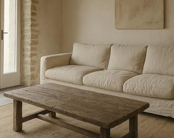 May include: A rustic, rectangular wooden coffee table with a distressed finish sits on a woven rug. A cream-colored sofa with three cushions is behind the table. The room has a neutral color palette.