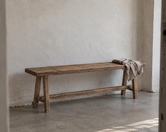 May include: A long, rectangular wooden bench with a weathered, natural brown finish. The bench features four sturdy, angled legs connected by a horizontal support beam. A folded, light-coloured fabric cloth rests on the right side of the seat.