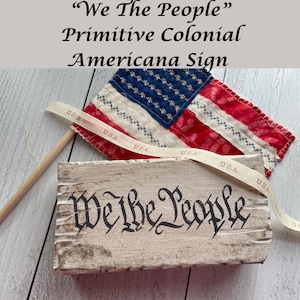 May include: A rustic wooden sign with the words "We The People" in black lettering. A small American flag and a ribbon with "USA" printed on it are also present, evoking a patriotic theme.