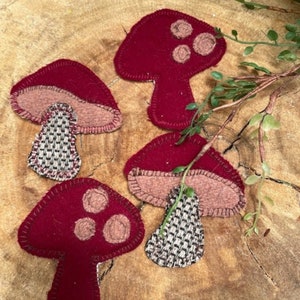 May include: Four felt mushroom ornaments in shades of red and brown. The mushrooms have a stitched detail around the edges and a textured fabric for the stem.