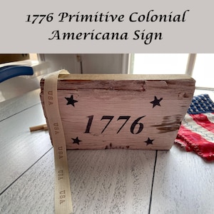 May include: A rustic wooden sign with the text "1776" and black star accents. The sign has a distressed, aged appearance with a cream-colored background and a ribbon that says "USA". The sign is a decorative item with a patriotic theme.