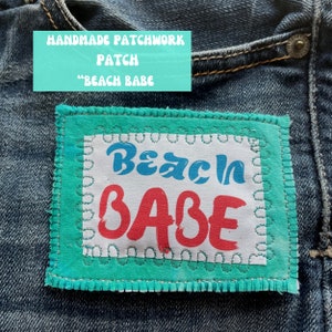 May include: A turquoise blue fabric patch with white stitching around the edges. The patch has a white background with the text "Beach Babe" in blue and red lettering.