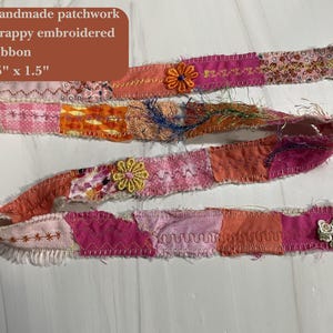 May include: Handmade patchwork ribbon, 45" x 1.5", in shades of pink, orange, and white. The ribbon is made of scrappy embroidered fabric and features decorative floral accents.