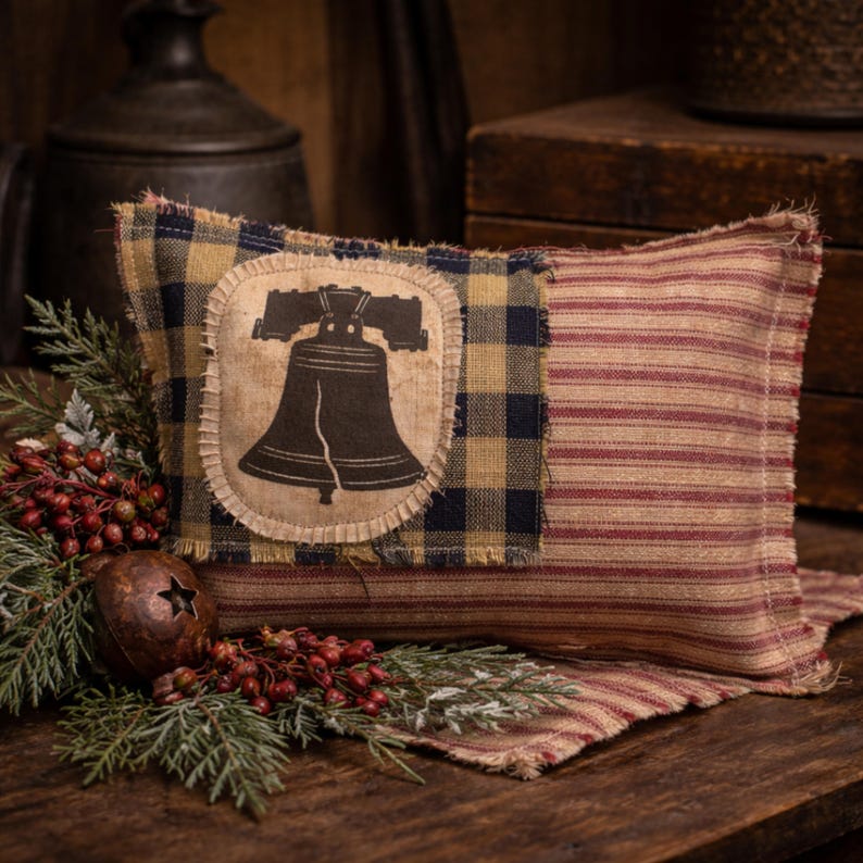 Op de afbeelding: Rechthoekig decoratief kussen met een patriottisch thema. Het kussen heeft een patchwork-ontwerp met een Liberty Bell-afbeelding, geruite en rood gestreepte stof. Het is geaccentueerd met nep groen en bessen.