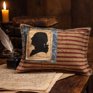 Op de afbeelding: Een rechthoekig kussen met een patriottisch thema. Het heeft een zwart silhouet van een historische figuur op een bruine achtergrond, tegen een blauwe bloemenstof. De hoofdbekleding van het kussen heeft rood-beige strepen.