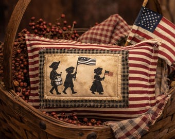 May include: A decorative pillow with a patriotic theme, featuring a black and white silhouette of children marching with flags and a drum. The pillow is red and white striped with a black and white checkered border, nestled in a woven basket with a small American flag.