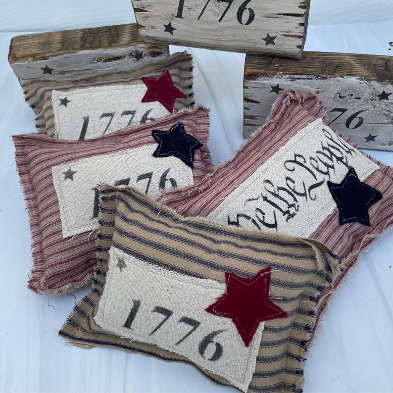 May include: Four decorative pillows with red, white, and blue striped fabric. The pillows feature the year 1776 and the phrase "We the People" in black lettering. The pillows are decorated with red, blue, and black stars.