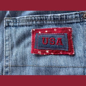 May include: A close-up of a blue denim patch with red stitching and white stars. The patch has the word "USA" embroidered in red thread.