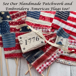 May include: Handmade patchwork and embroidered American flags in red, white, and blue. The flags feature stars and stripes, with some flags mounted on wooden dowels. A wooden block with "1776" is also present.