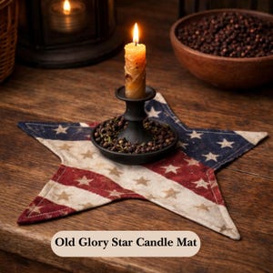May include: A lit candle in a black candle holder sits on a star-shaped candle mat. The mat features a distressed American flag design with red and blue stripes and white stars. The text "Old Glory Star Candle Mat" is at the bottom.