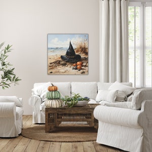 May include: A black witch's hat sits on a sandy beach with three small pumpkins. The hat has a pointed top and a wide brim. The pumpkins are orange and green. The background is a blue sky with white clouds.