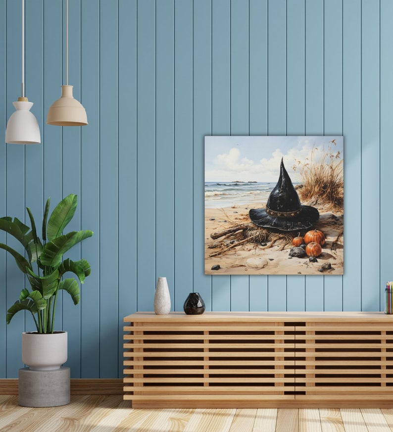 May include: A black witch's hat with a wide brim sits on a sandy beach with three small pumpkins. The hat is made of felt and has a pointed crown. The pumpkins are orange and have a stem on top. The background is a blue sky with white clouds.