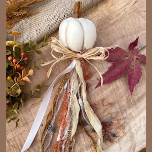 May include: A white felt pumpkin with a brown stem and a bow made of natural twine. The pumpkin is attached to a long strand of frayed fabric in shades of white, brown, and orange.