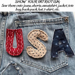May include: Close-up of denim jeans with the letters "USA" appliqued on the back pocket. The "U" is red with a white leaf pattern, the "S" is tan with black stars, and the "A" is navy blue with white stars. Text above reads "SHOW YOUR PATRIOTISM".