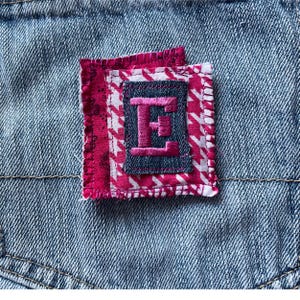 May include: A pink and white checkered fabric patch with a pink letter E sewn on top. The patch is attached to a blue denim fabric.