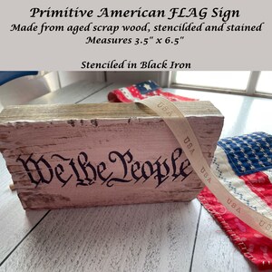 May include: A rustic, rectangular sign made from aged wood, stenciled with "We the People" in black iron. The sign measures 3.5" x 6.5" and features a distressed, whitewashed finish. A ribbon with "USA" text is draped across the sign.