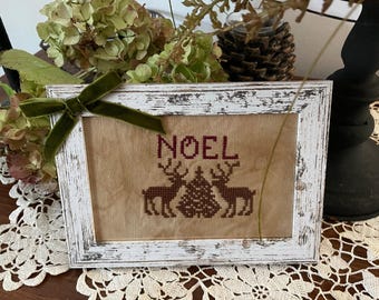 May include: A framed cross-stitch featuring the word "NOEL" in burgundy, with two reindeer flanking a Christmas tree. The frame is white with a distressed finish, and a green velvet ribbon is tied on the top left corner. The background is a neutral tan color.