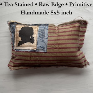 Op de afbeelding: Handgemaakt kussen met theekleurige stof en rafelige randen. Het kussen heeft een silhouet op een blauwe patch en rood-beige strepen. Het kussen meet ongeveer 20 x 13 cm.