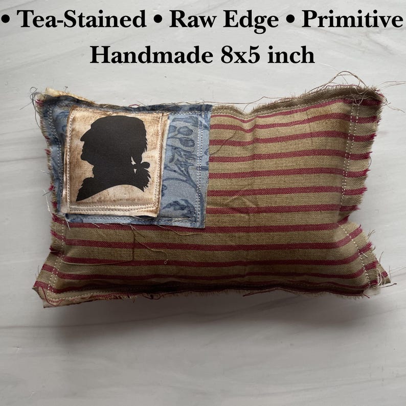 Op de afbeelding: Handgemaakt kussen met theekleurige stof en rafelige randen. Het kussen heeft een silhouet op een blauwe patch en rood-beige strepen. Het kussen meet ongeveer 20 x 13 cm.