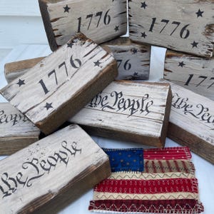 May include: A collection of distressed wooden blocks with the year "1776" and star accents painted in black. Some blocks feature the phrase "We the People" in a script font. A small, fabric American flag is also present, showcasing red, white, and blue colors.