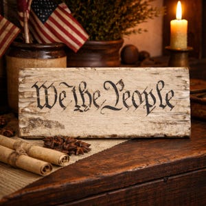 May include: A rustic wooden sign with the words "We the People" in black script. The sign is aged and distressed, with a weathered appearance. American flags and a lit candle are in the background, evoking a patriotic theme.