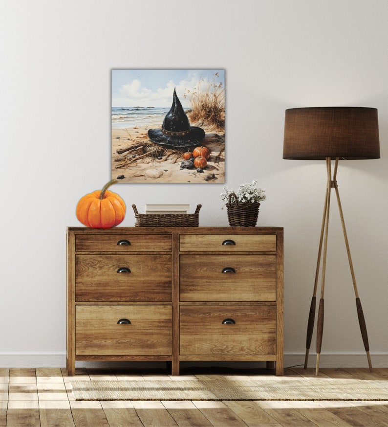 May include: A black witch's hat with a wide brim sits on a sandy beach with three small orange pumpkins. The hat is surrounded by dried grasses and driftwood. The beach is in the background with a blue sky and white clouds.